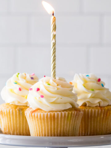 Four birthday cupcakes on a cake stand with a lit gold birthday candle in the front cupcake.