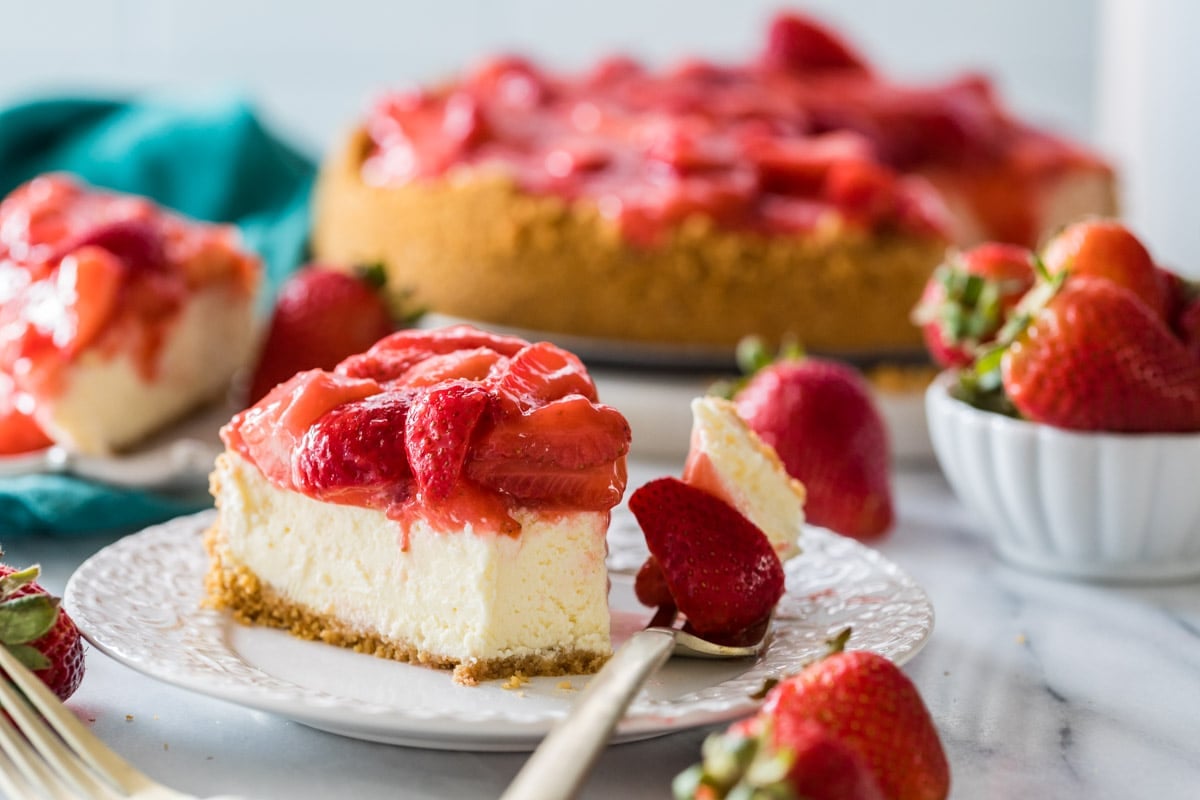 Forkful of cheesecake beside a slice of strawberry topped cheesecake on a white plate.