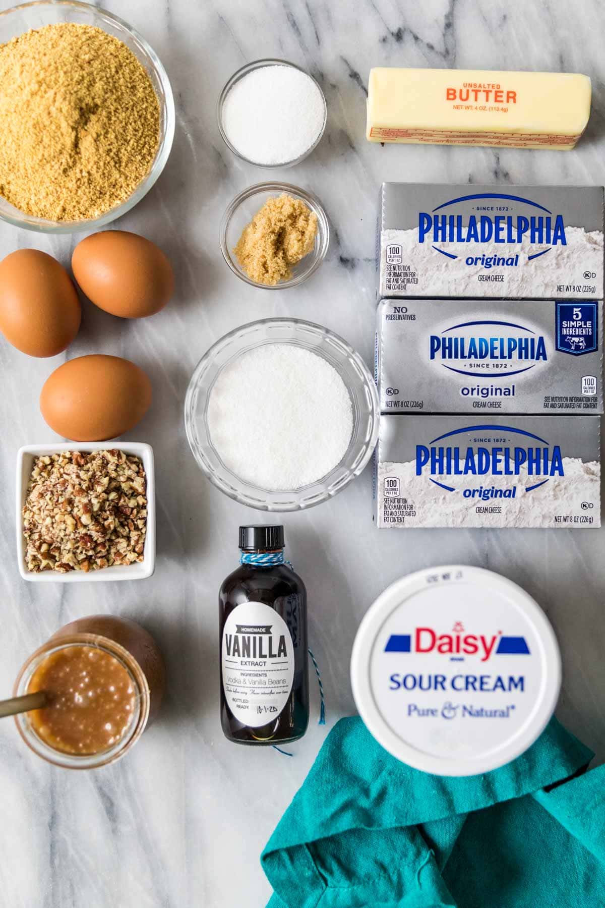 Overhead view of ingredients including graham cracker crumbs, pecans, cream cheese, caramel sauce, and more.