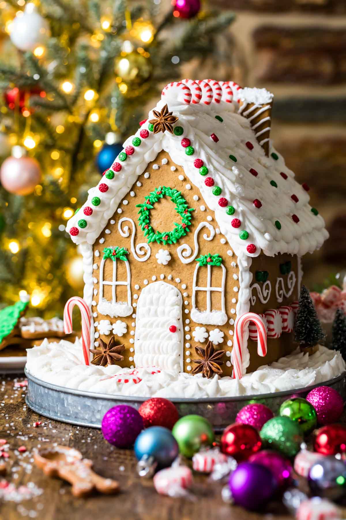 Intricately decorated gingerbread house on a metal plate with christmas ornaments in the foreground and a lit tree in the background.