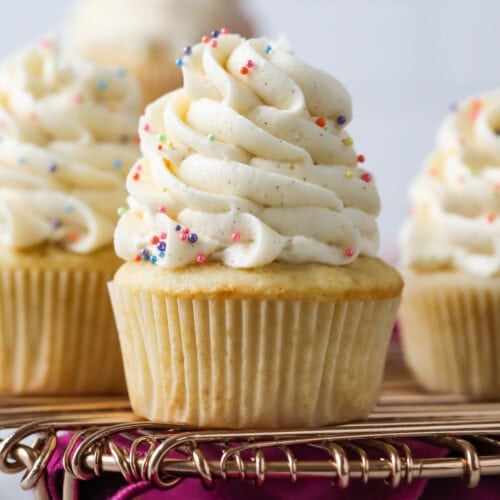 Vanilla cupcakes topped with tall swirls of vanilla bean buttercream on a cooling rack.