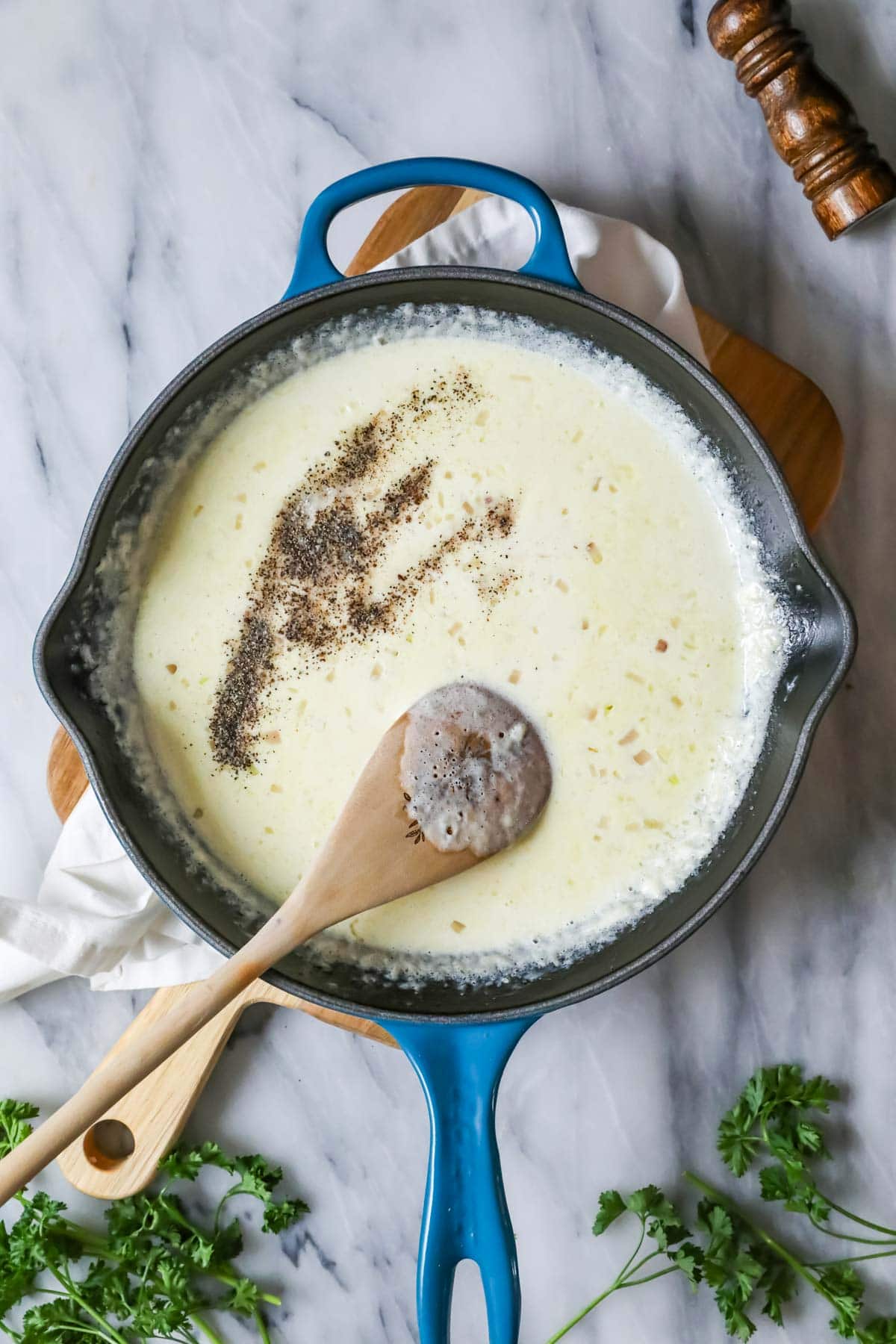 Spoon stirring black pepper, nutmeg, and salt into a cream sauce.