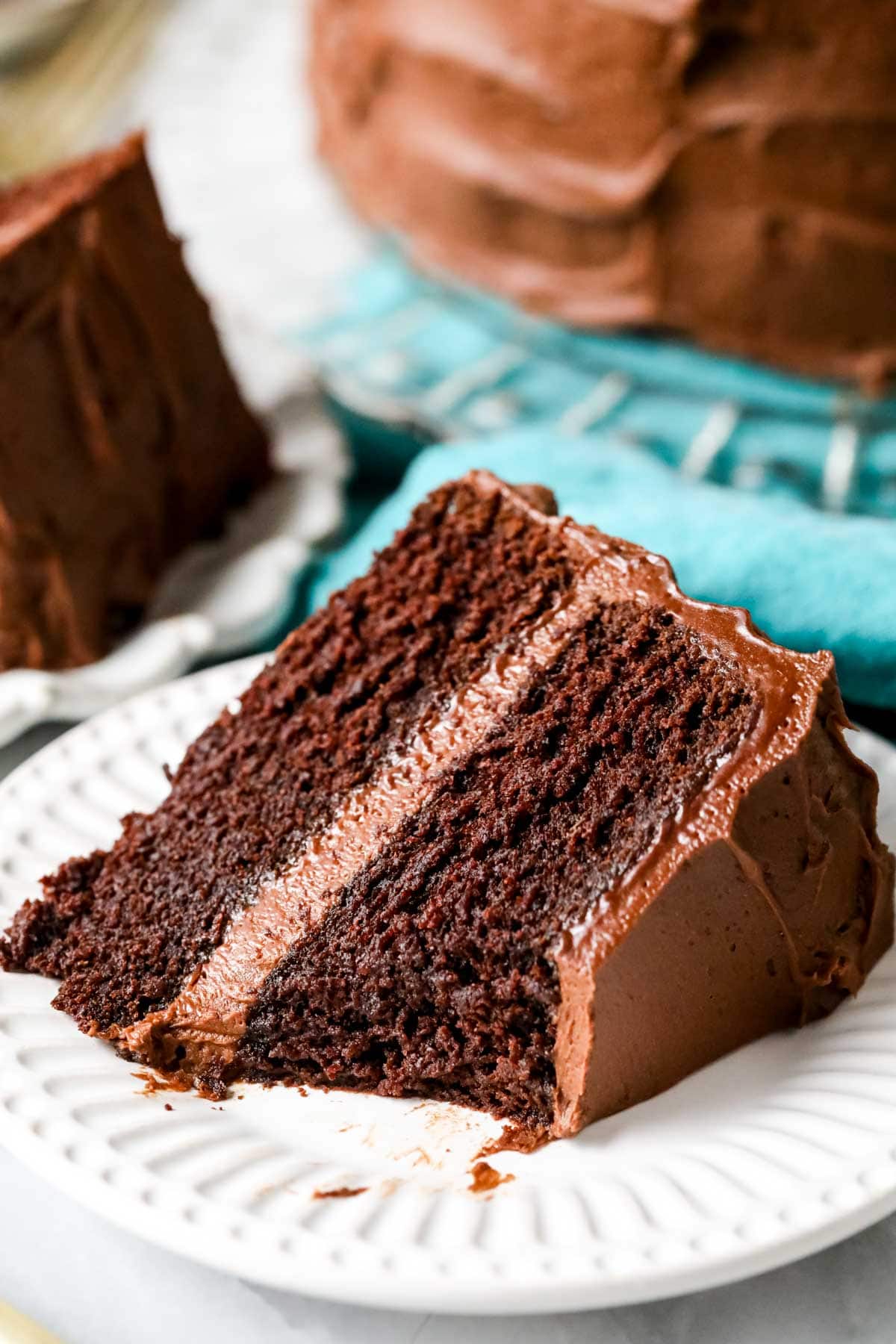 Slice of fudgy chocolate cake on a white plate with cake on crystal platter over blue cloth in background.