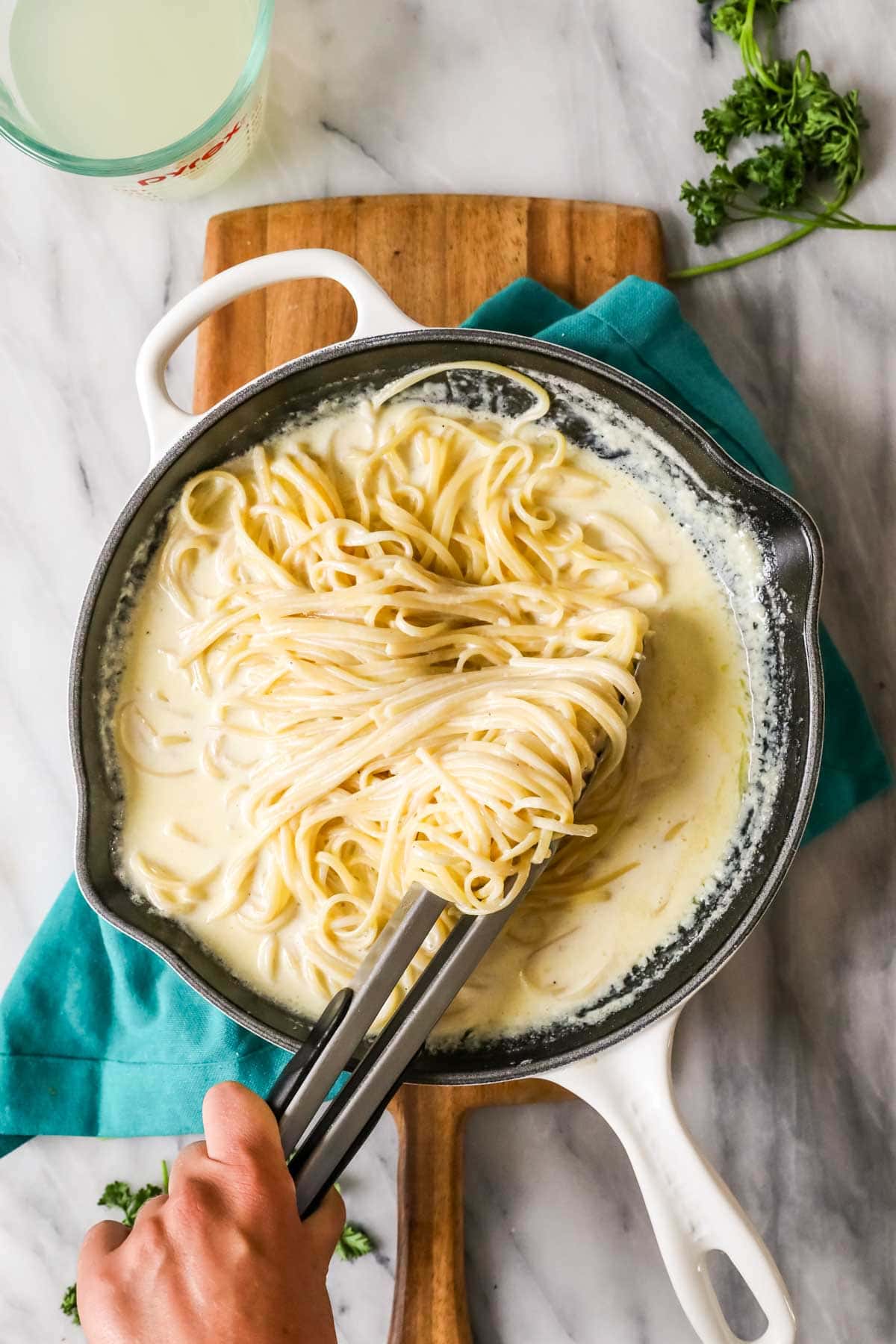 Overhead view of long noodles being tossed into alfredo sauce using tongs.