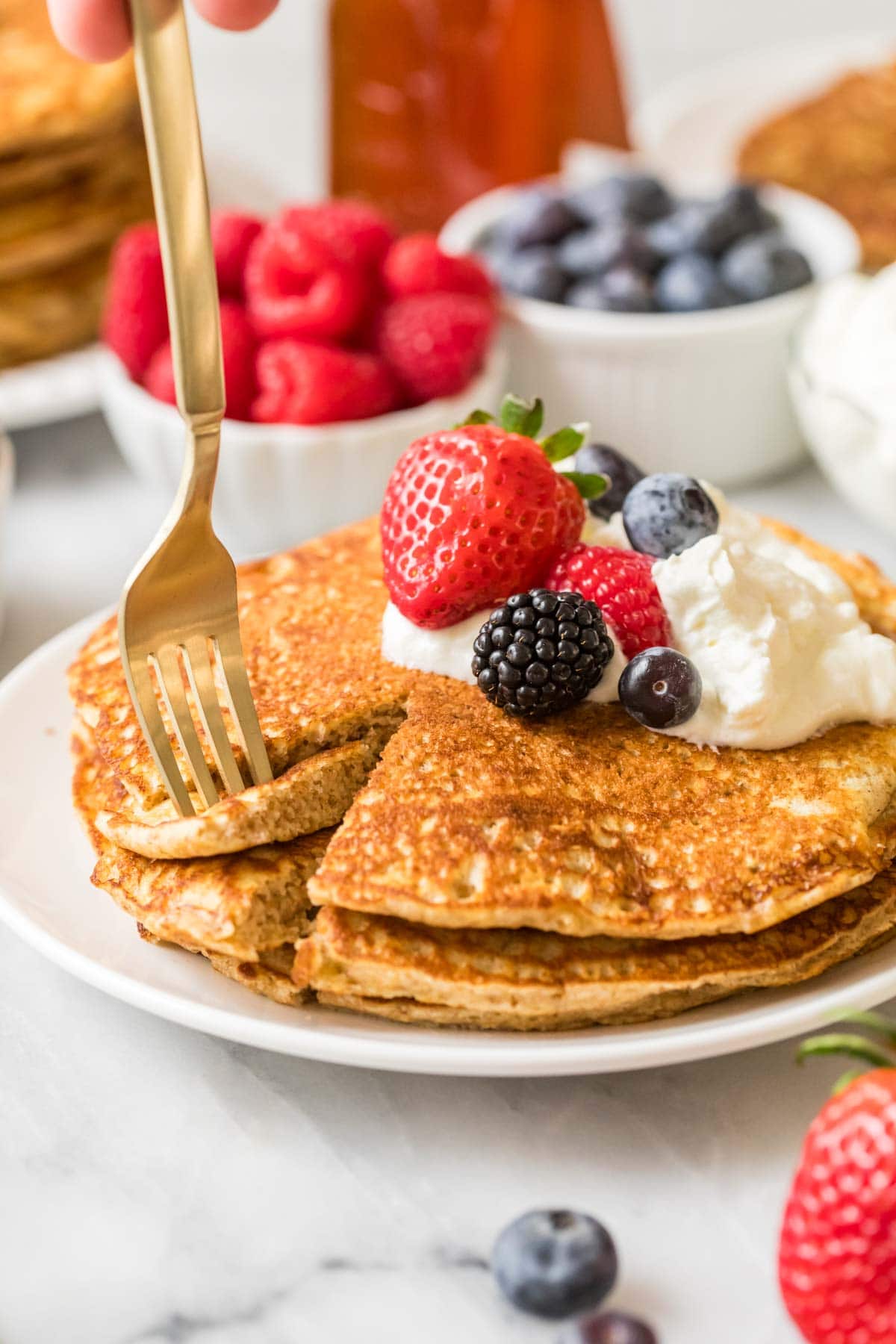 Fork taking a bite out of a stack of whole wheat pancakes topped with whipped cream and berries.
