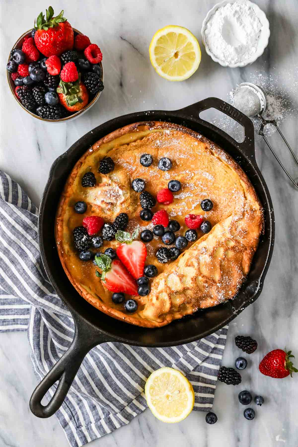 Overhead view of a Dutch baby topped with fresh berries and powdered sugar in a cast iron skillet.