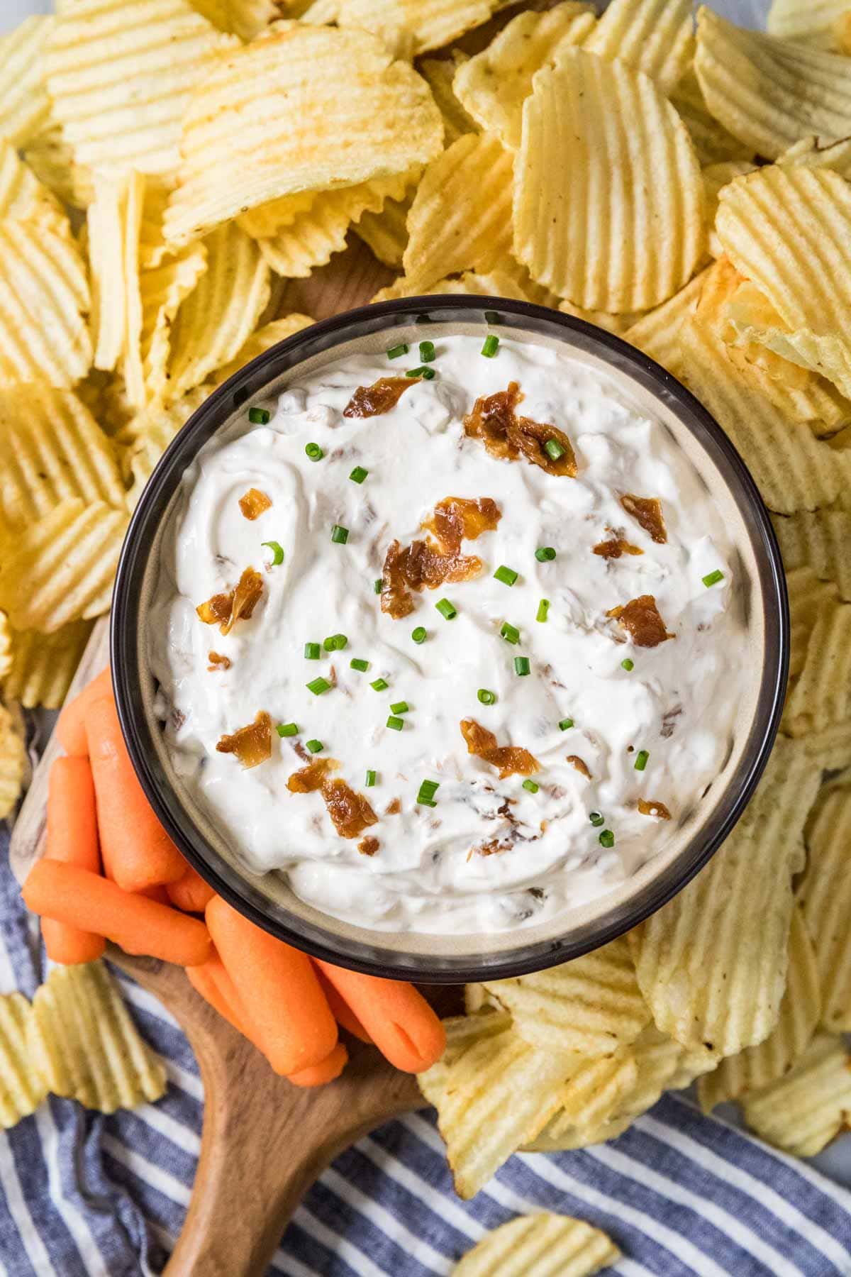 Overhead view of a bowl of french onion dip surrounded by ripple potato chips and baby carrots.