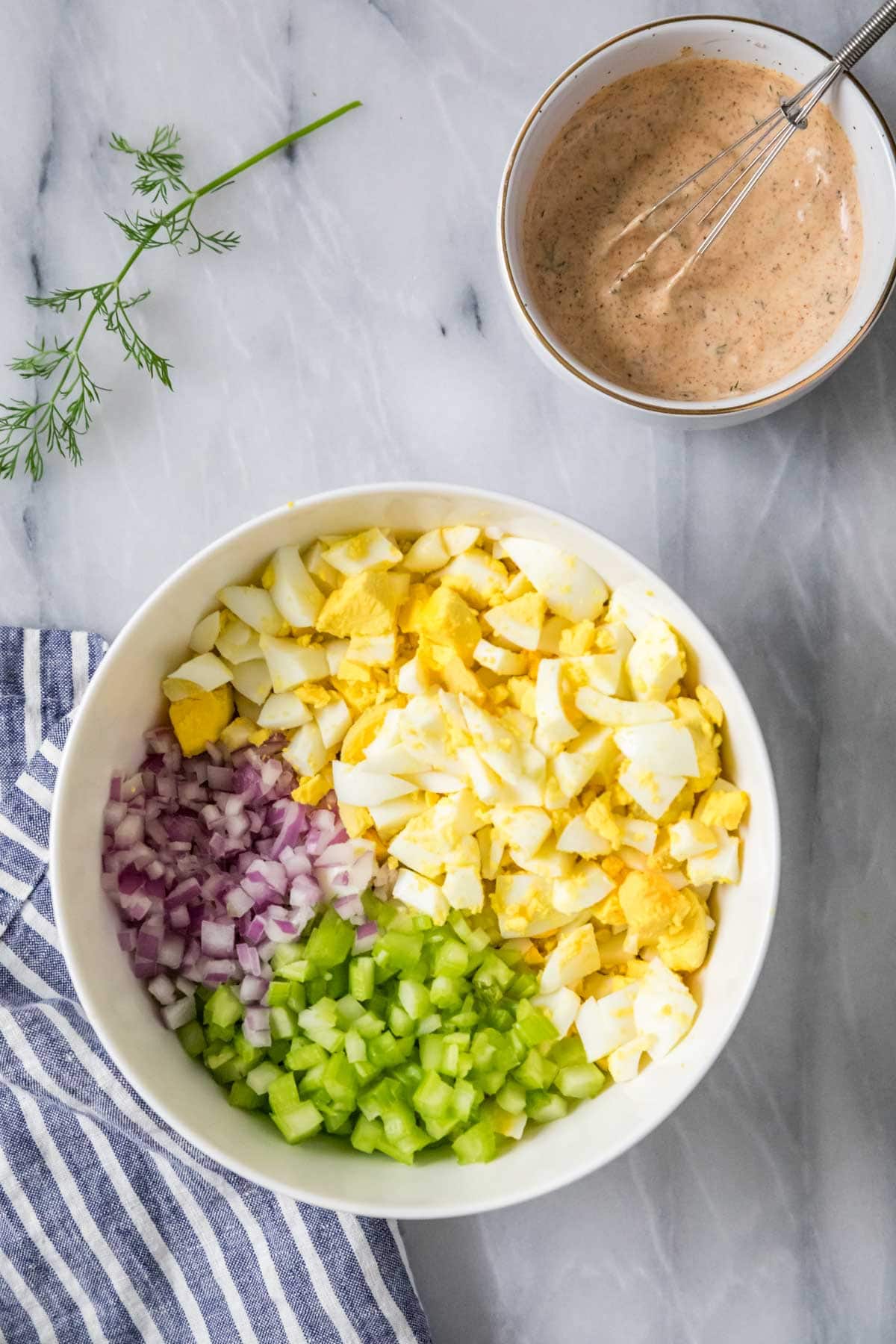 Overhead view of a serving bowl with chopped eggs, onion, and celery next to a bowl of dressing.