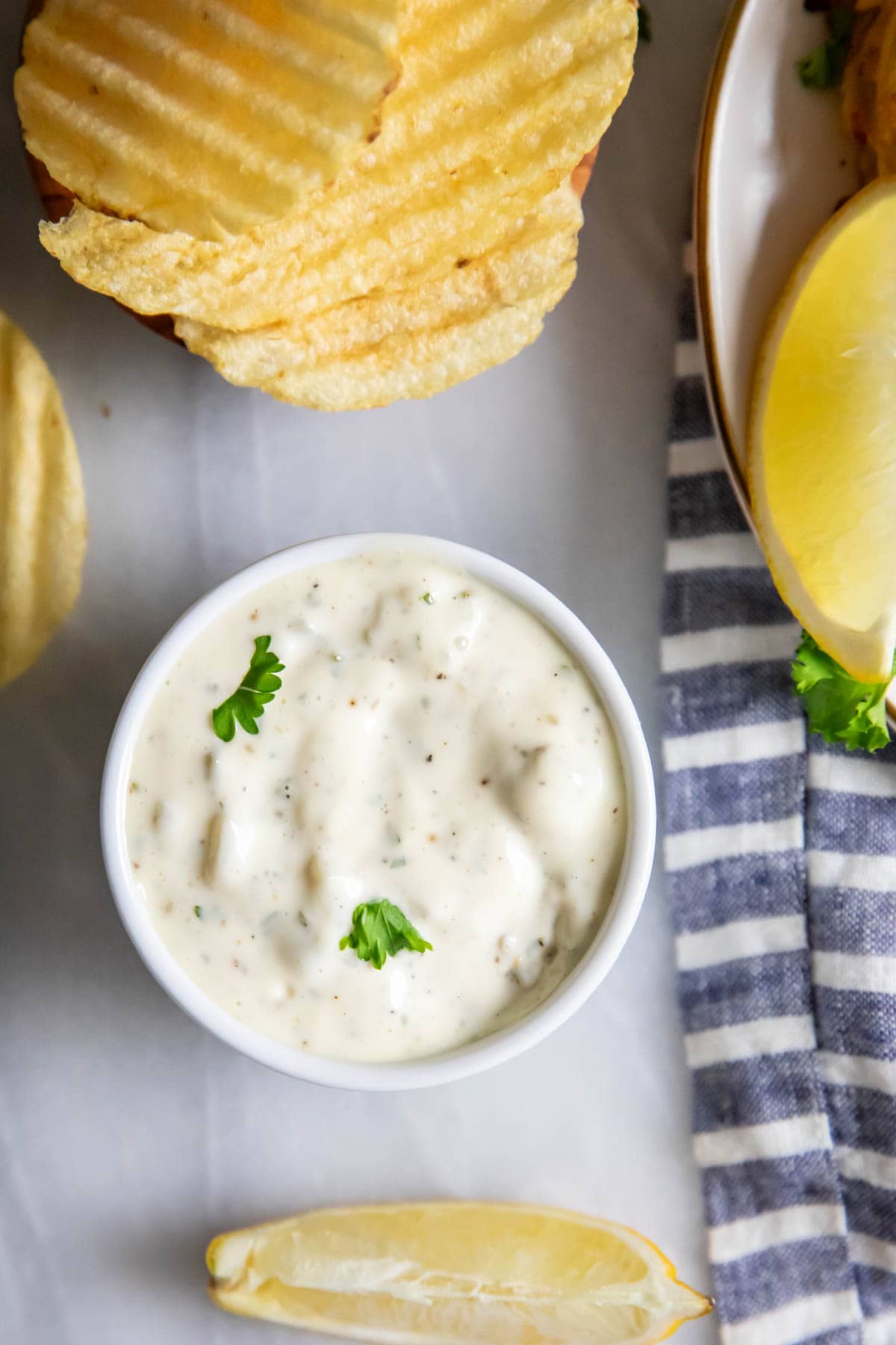 Overhead view of a bowl of homemade tartar sauce surrounded by potato chips and lemon wedges.
