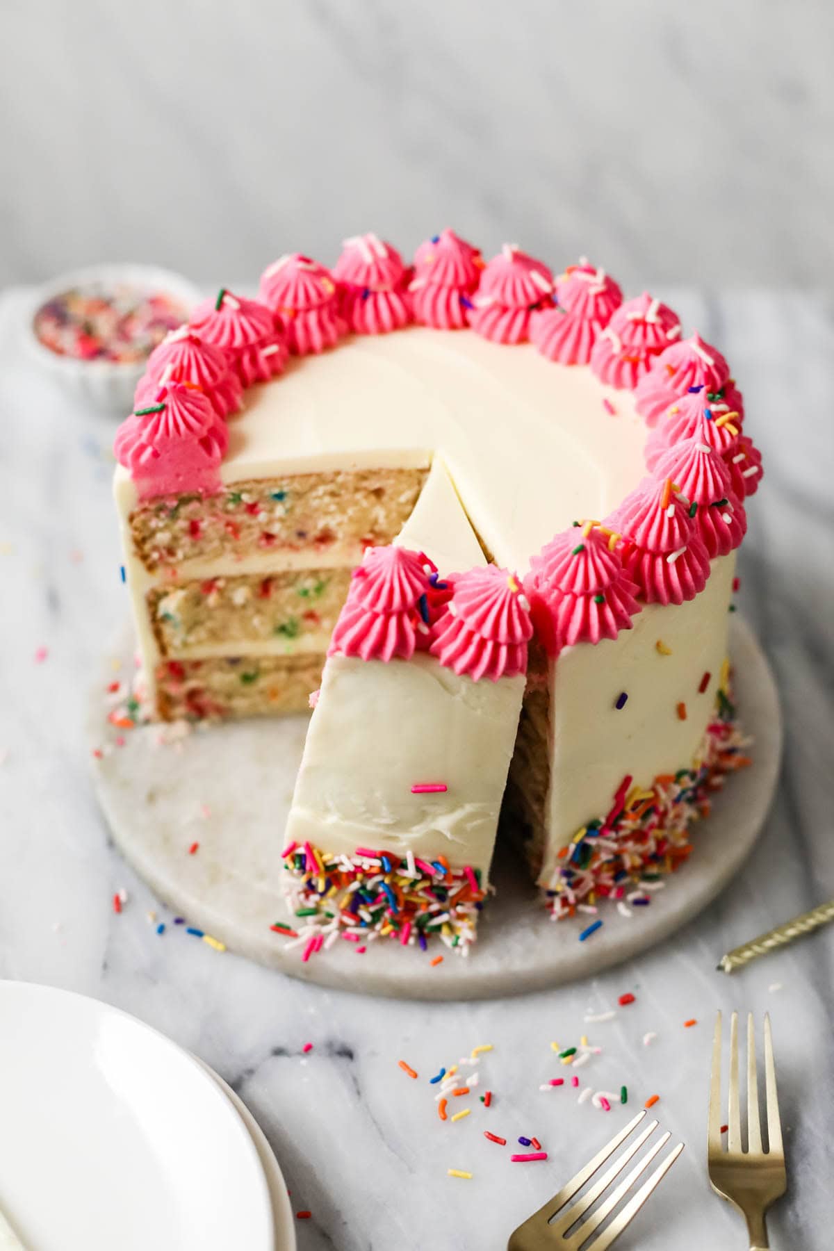 Tall funfetti cake decorated with a bright pink frosting border and sprinkles with a few slices cut from it.