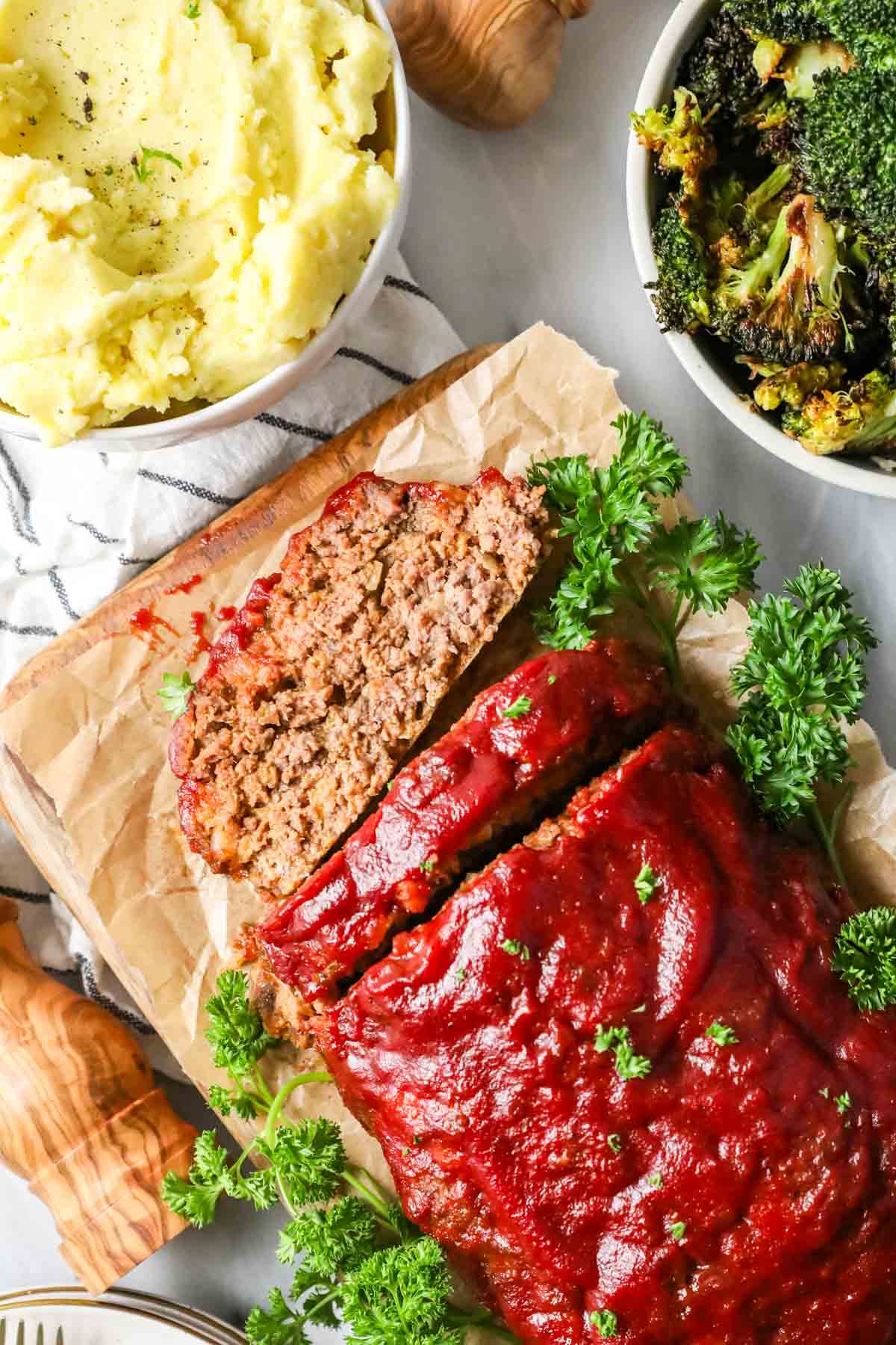 Overhead view of a meatloaf with two slices cut surrounded by bowls of mashed potatoes and roasted broccoli.