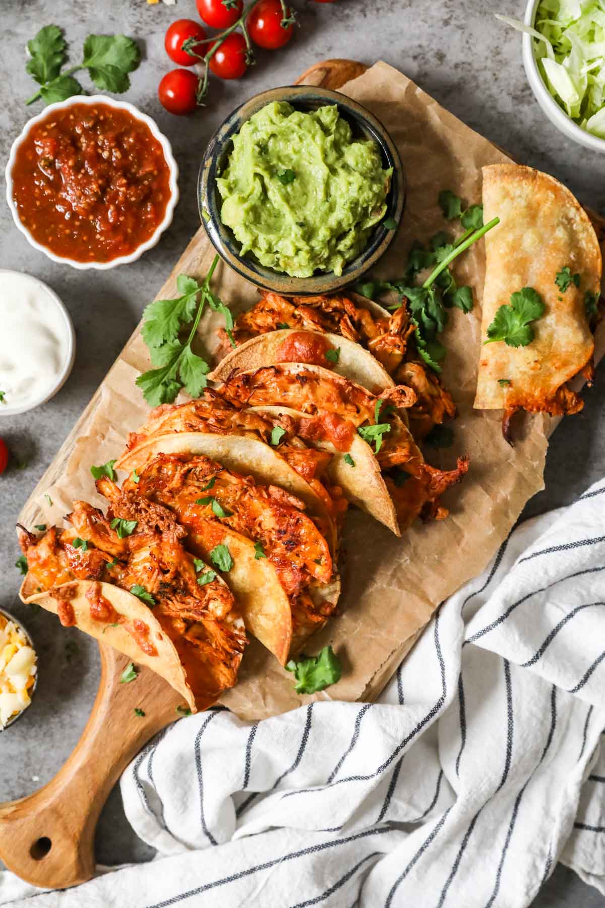 Overhead view of crispy chicken tacos on a wood cutting board with sides of guacamole, salsa, sour cream, and cheese.