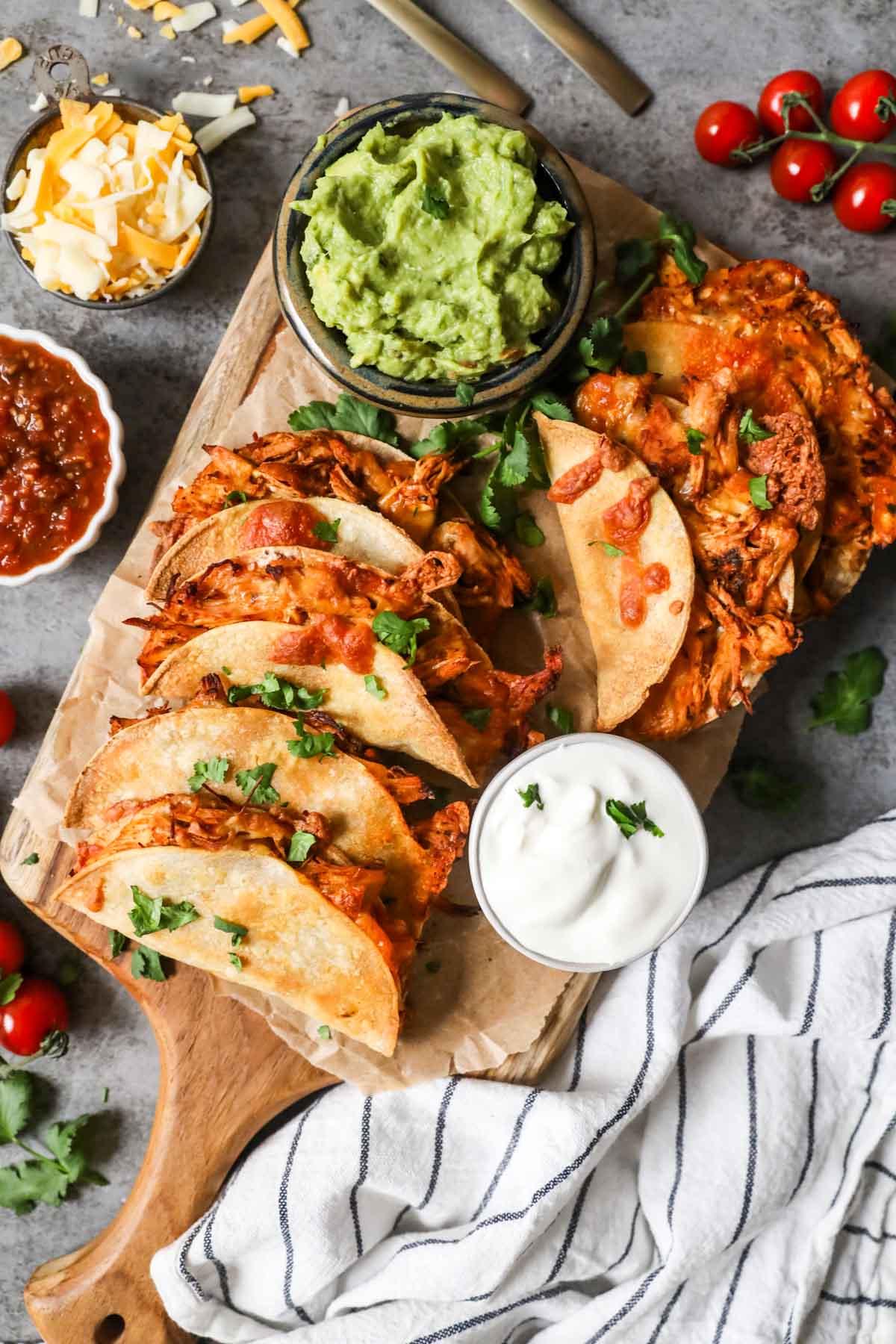Overhead view of crispy chicken tacos on a wood cutting board with sides of guacamole, salsa, sour cream, and cheese.