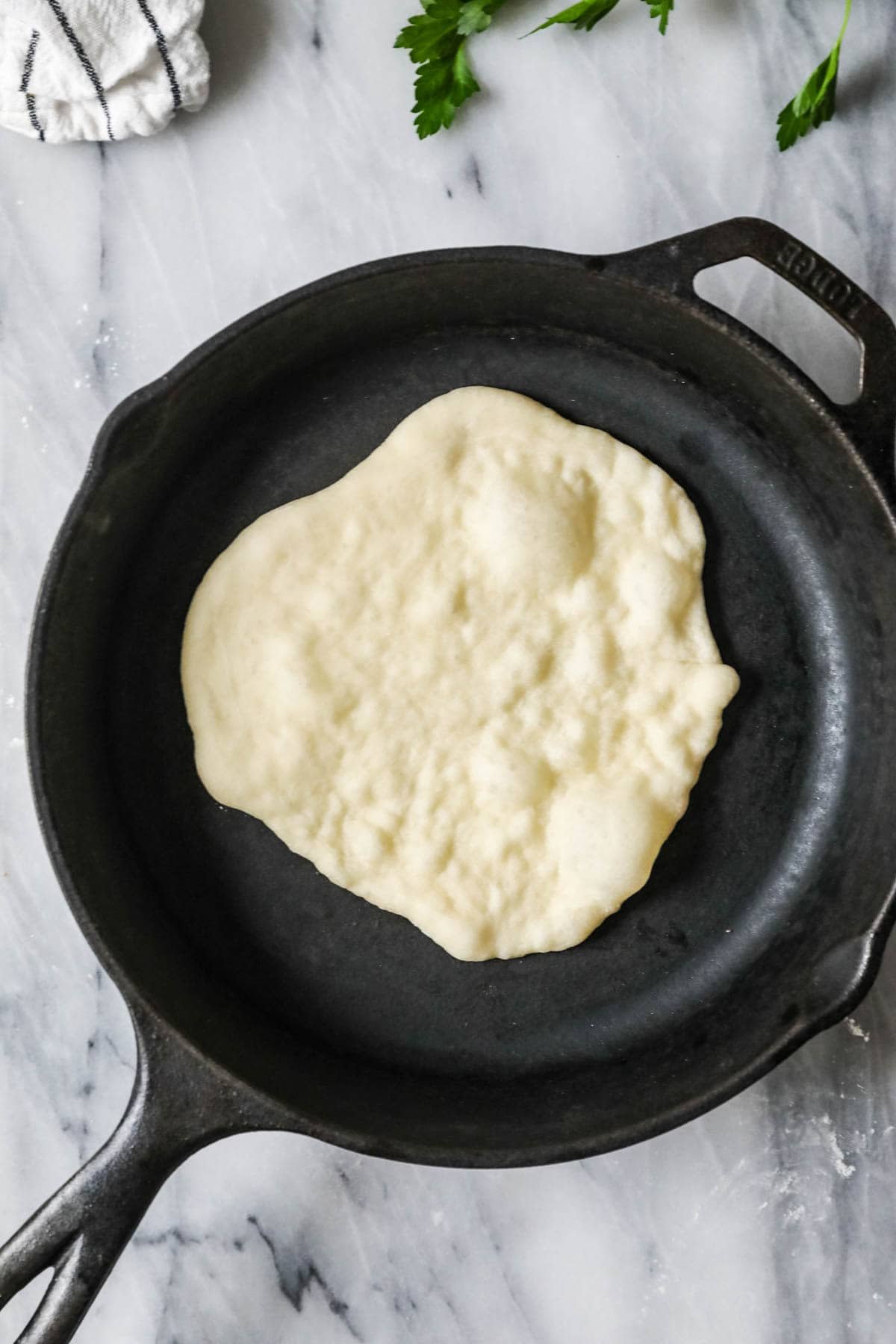 Overhead view of naan cooking in a cast iron pan with large bubbles forming on the surface.
