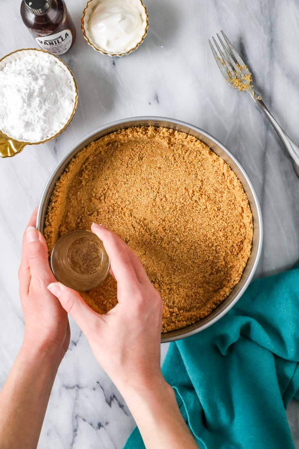 Overhead view of a graham cracker crust being pressed into a springform pan with a glass.