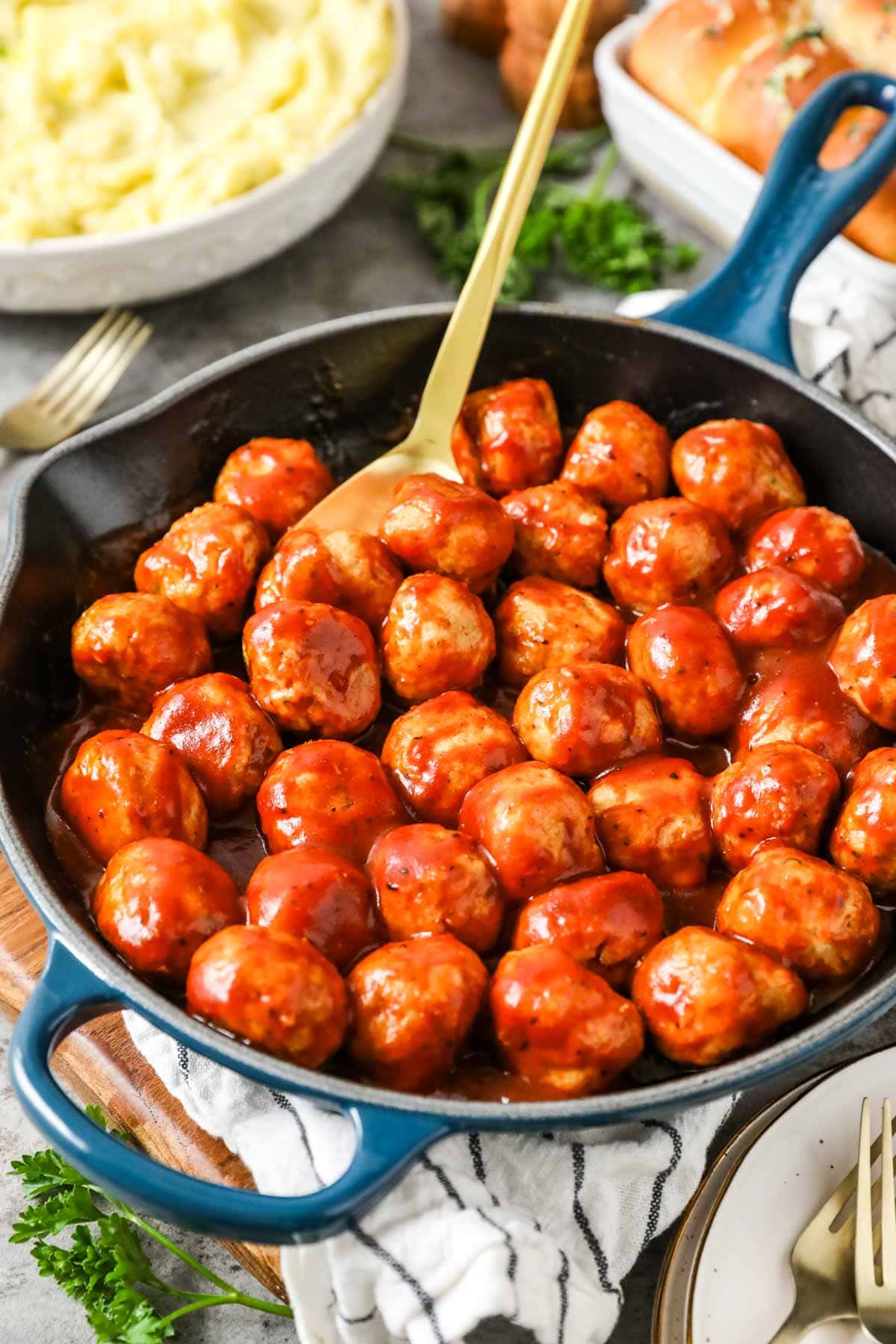 Black skillet with a blue handle containing freshly made BBQ chicken meatballs with a gold serving spoon sticking out.