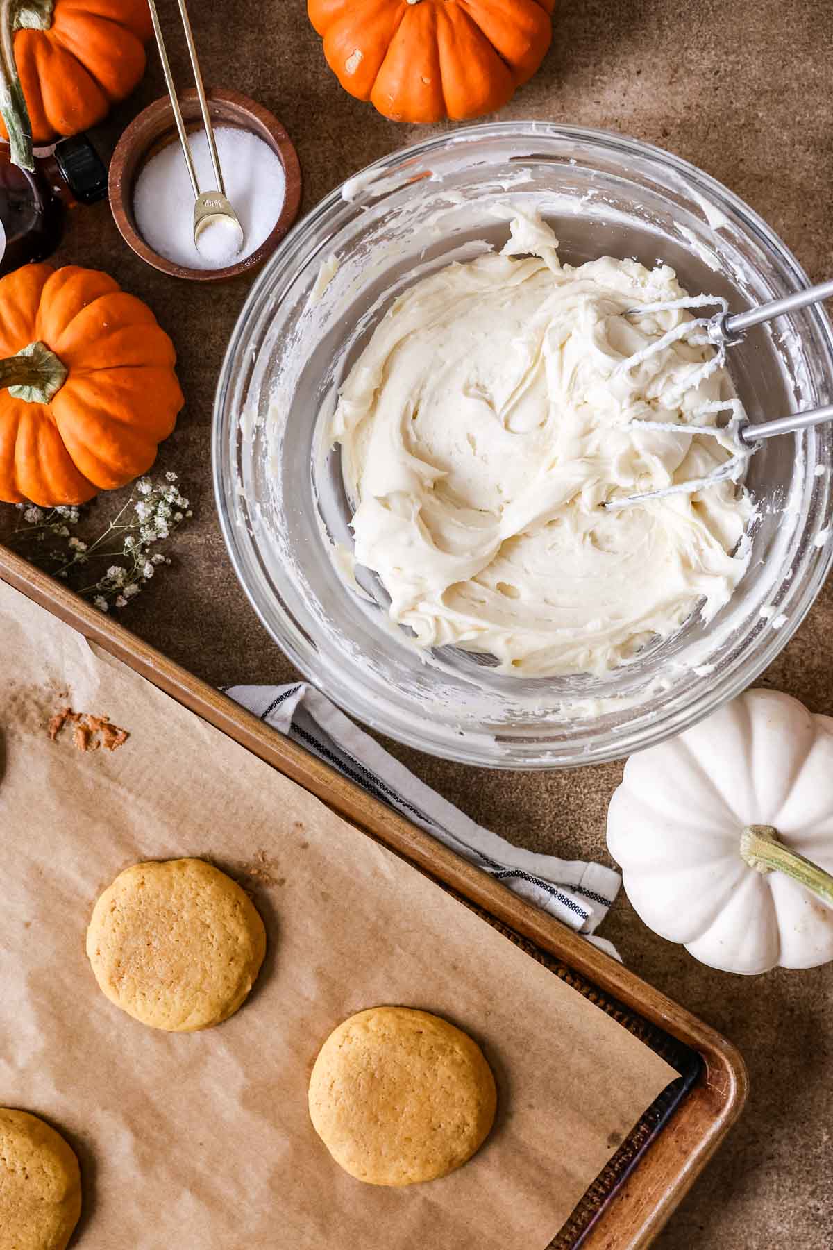 Mixed cream cheese frosting with pumpkin cookies on baking sheet beside it, pumpkins around the bowl.