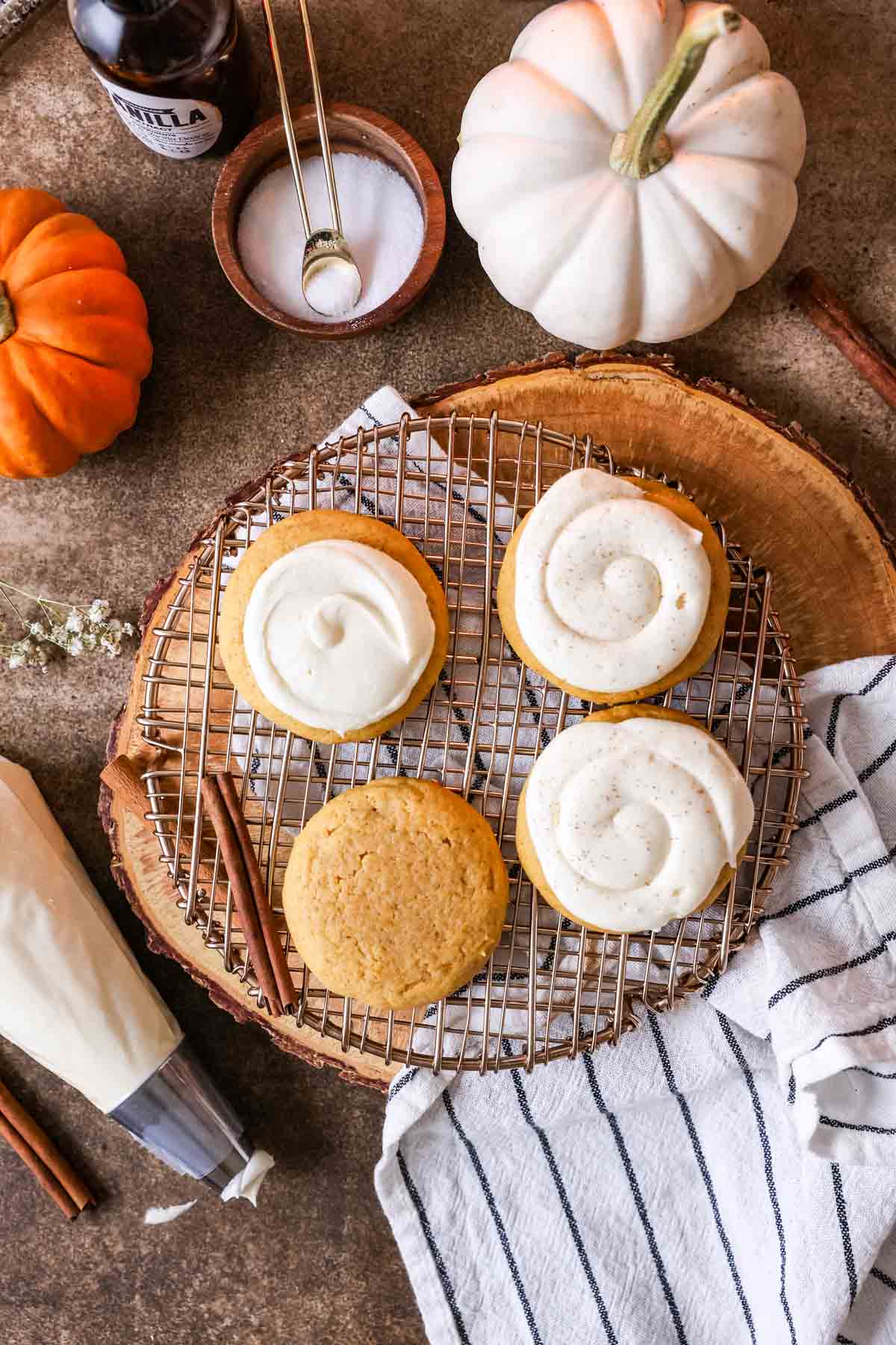 Overhead view of pumpkin cookies with cream cheese frosting, one cookie not yet frosted, pumpkins, salt, vanilla and frosting bag in photo.